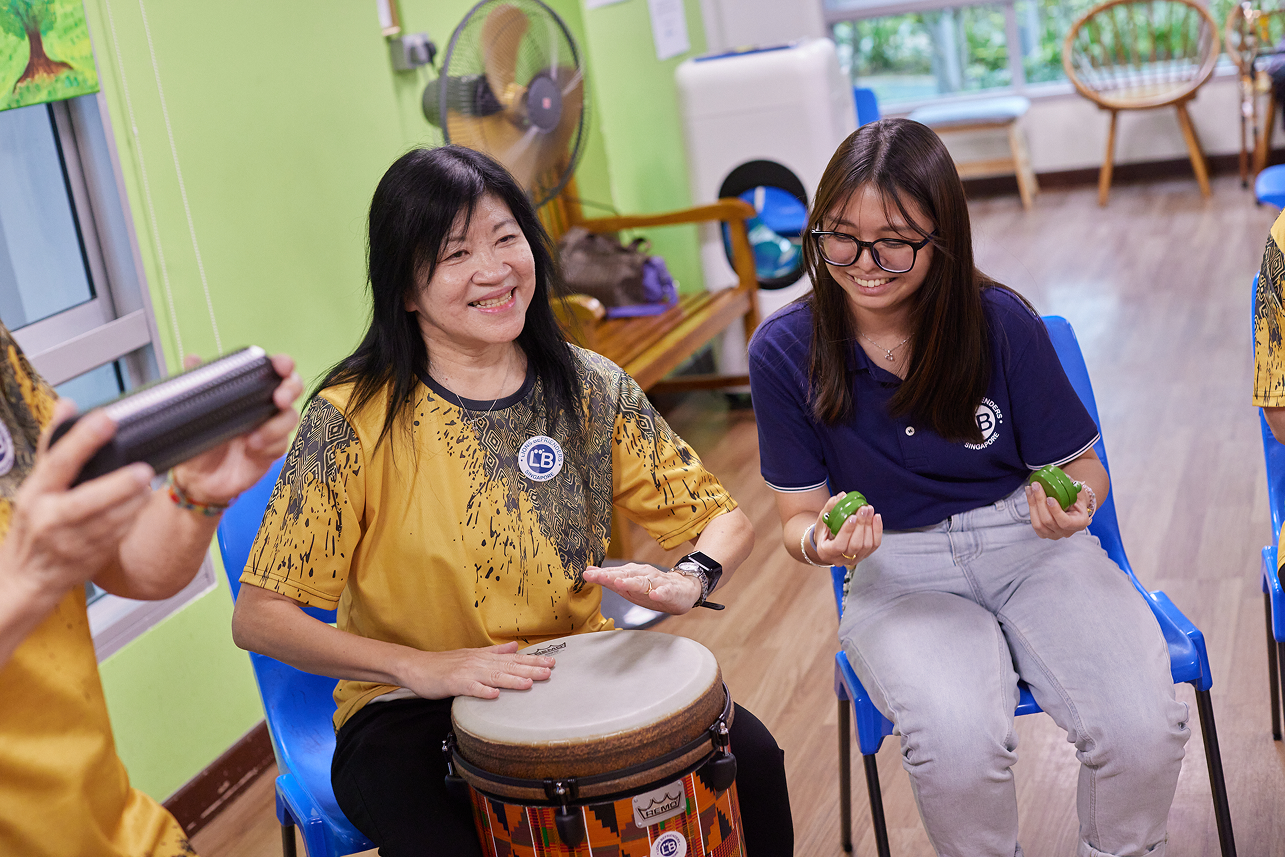 Two women playing musical instruments, wearing shirts with "LB" logo, seated on blue chairs.